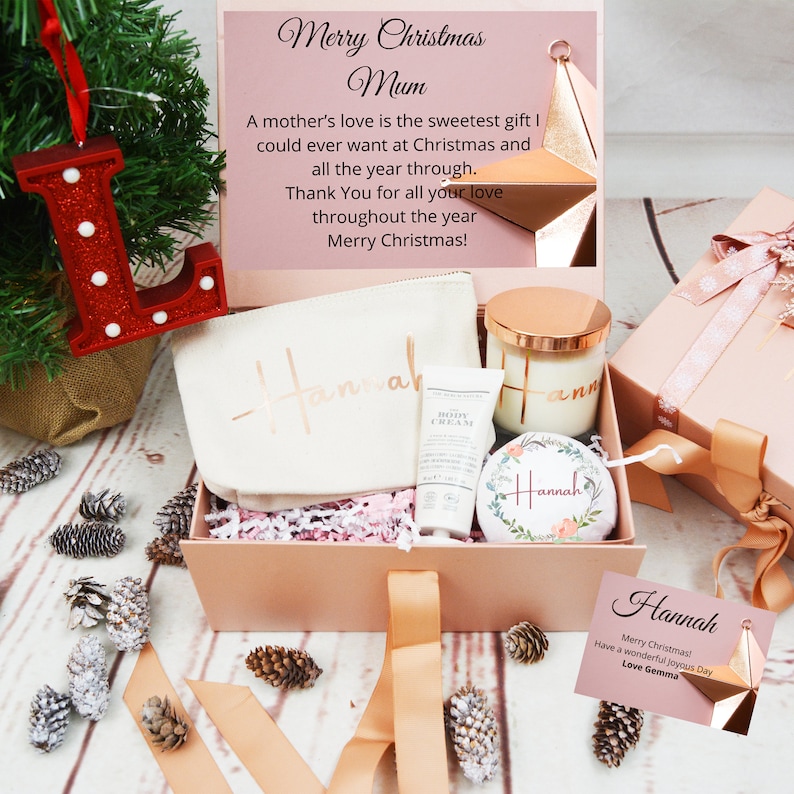 May include: A pink gift box with a white canvas bag, a small bottle of body cream, a candle, and a floral ornament. The box is decorated with a gold ribbon and the words "Merry Christmas Mum" are printed on the top of the box. The ornament has the name "Hannah" written on it.