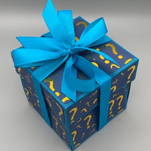 May include: A blue gift box with a blue ribbon and gold question marks printed on the box.