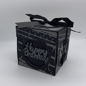 May include: A square black and grey gift box featuring a tyre tread pattern on the sides and a black ribbon bow on top. The front panel displays a circular cutout with the text Happy Birthday in black script lettering against a dark background.