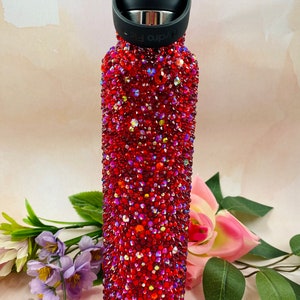 Op de afbeelding: Een rode Hydro Flask waterfles bedekt met glinsterende rode strass-steentjes. De fles heeft een zwarte dop en handvat. De fles wordt omringd door bloemen en bladeren.
