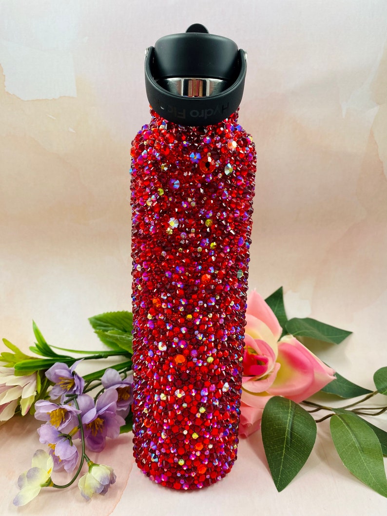 Op de afbeelding: Een rode Hydro Flask waterfles bedekt met glinsterende rode strass-steentjes. De fles heeft een zwarte dop en handvat. De fles wordt omringd door bloemen en bladeren.