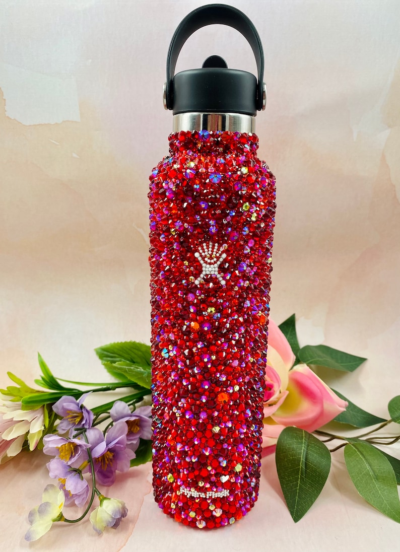 Op de afbeelding: Een rode waterfles bedekt met glinsterende rode strass-steentjes. De fles heeft een zwarte handgreep en deksel, met een zilveren rand. De fles is versierd met een wit logo. De fles is omgeven door bloemen.