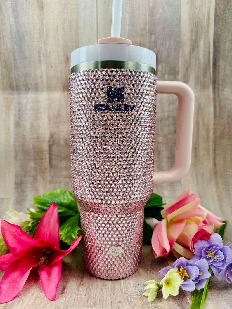 Pode incluir: Um copo de a&ccedil;o inoxid&aacute;vel rosa com al&ccedil;a e tampa transparente. O copo &eacute; coberto com strass rosa e tem a palavra "Stanley" impressa no lado. O copo est&aacute; sobre uma superf&iacute;cie de madeira com flores.