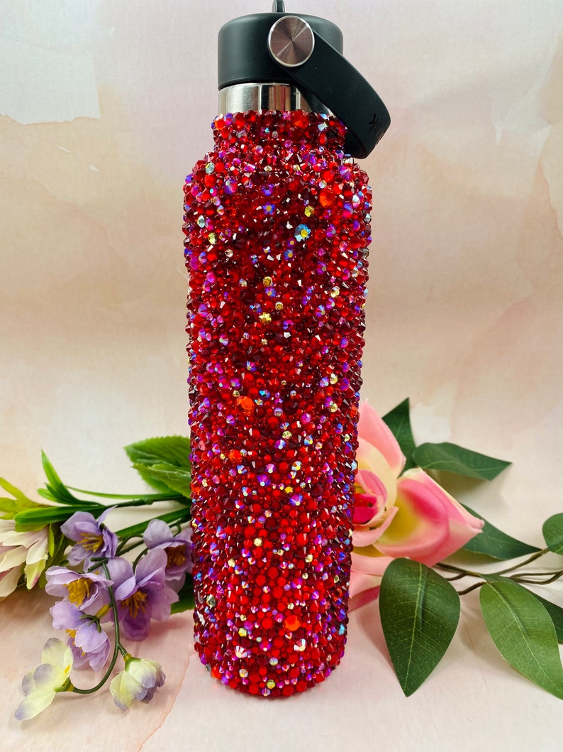 Op de afbeelding: Een rode waterfles bedekt met glinsterende rode strass-steentjes. Het heeft een zwarte dop en handvat. De achtergrond toont bloemen en bladeren. De fles is ongeveer 25 cm hoog.