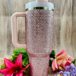 Pode incluir: Um copo de a&ccedil;o inoxid&aacute;vel rosa com uma tampa transparente e uma al&ccedil;a rosa. O copo &eacute; coberto com strass.