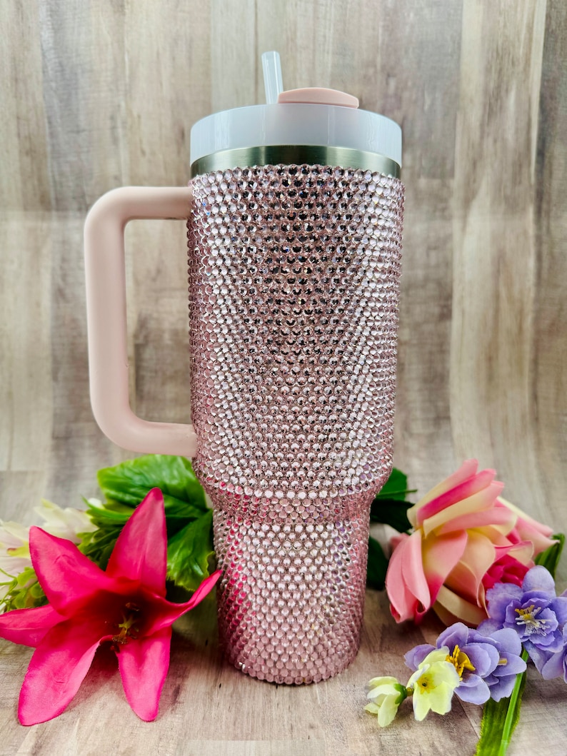Pode incluir: Um copo de a&ccedil;o inoxid&aacute;vel rosa com uma tampa transparente e uma al&ccedil;a rosa. O copo &eacute; coberto com strass.