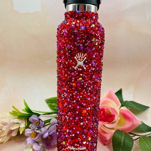 Op de afbeelding: Een rode waterfles bedekt met glinsterende rode strass-steentjes. Het heeft een zwarte dop en een zilveren rand. Het merklogo is zichtbaar. De fles is omringd door bloemen.