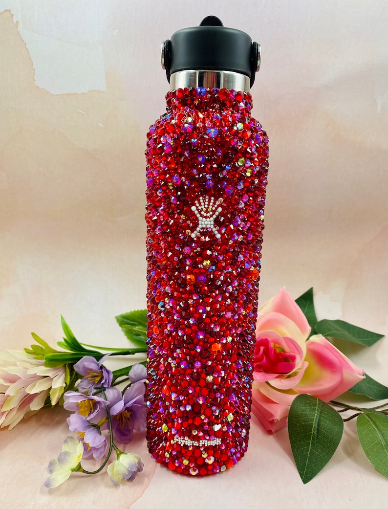 Op de afbeelding: Een rode waterfles bedekt met glinsterende rode strass-steentjes. Het heeft een zwarte dop en een zilveren rand. Het merklogo is zichtbaar. De fles is omringd door bloemen.