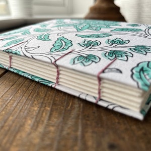 May include: A white journal with a teal floral pattern and pink stitching. The journal is closed and lying on a wooden surface.