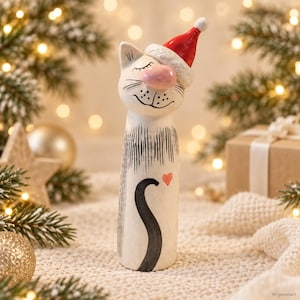 Könnte beinhalten: Eine hohe, zylindrische weiße Katzenfigur mit glatter Oberfläche, die eine rot-weiße Weihnachtsmütze trägt. Die Katze hat eine rosa Nase, geschlossene Augen, schwarze Schnurrhaare, einen aufgemalten schwarzen Schwanz und ein kleines rotes Herz.