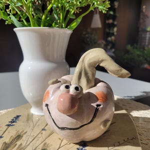 Könnte beinhalten: Eine hellrosa Kürbisfigur aus Keramik mit glänzender Oberfläche, die eine große runde Nase, zwei kleine Augen, orange bemalte Wangen und ein breites, schwarz gemaltes Lächeln aufweist. Der Kürbis hat einen geschwungenen, hellbeigen Stiel.