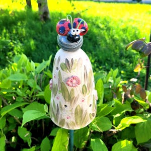 Könnte beinhalten: Ein Keramik-Gartenstecker mit glockenförmigem Körper auf cremefarbenem Grund, bemalt mit grünen Blättern und kleinen rosa Blüten. Oben sitzt eine Marienkäferfigur mit roten Flügeln und schwarzen Punkten auf einem grauen Kopf mit zwei weißen Augen.