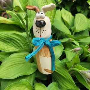 Puede incluir: Una figura de perro de cerámica con cuerpo color crema, una oreja marrón y nariz negra. El perro tiene ojos grandes y redondos, una boca pequeña roja y una cinta de tela color verde azulado atada al cuello. El cuerpo tiene marcas marrones pintadas.