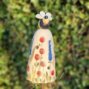 Könnte beinhalten: Ein skurriler Gartenstecker mit einer Keramikbiene auf einer glockenförmigen Form. Die Glocke ist mit roten Blumen, blauen Akzenten und kleinen Bienenillustrationen verziert. Der Stecker ist aus Metall und für den Außenbereich bestimmt.