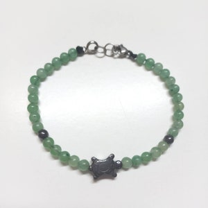 May include: A bracelet made of small, round, light green beads. A dark grey turtle charm is the focal point. The bracelet has a silver-coloured clasp and connecting hardware.