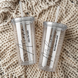 Könnte beinhalten: Zwei durchsichtige Acrylbecher mit passenden Deckeln und geraden Trinkhalmen. Jeder Becher ist mit schwarzem Text versehen, einem Namen in Schreibschrift und einem Titel in serifenloser Schrift. Einer zeigt Ariana the Bride, der andere Melina Bridesmaid.