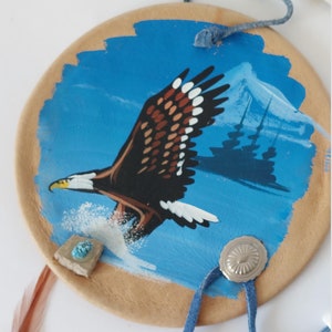 Peut inclure: Un attrape-rêves rond en cuir brun avec un fond bleu et une image peinte d'un aigle en vol. L'attrape-rêves a un cordon en cuir bleu et des perles blanches. Il y a une pierre turquoise et une conque argentée sur l'attrape-rêves.