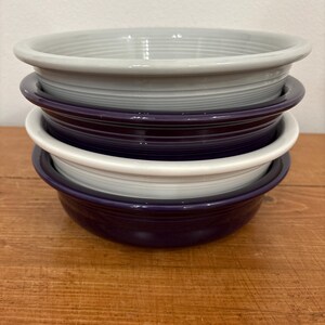 May include: A stack of four ceramic bowls in various colors: gray, purple, white, and dark purple. The bowls have a simple, rounded design with a slightly flared rim. They are stacked on a wooden surface.
