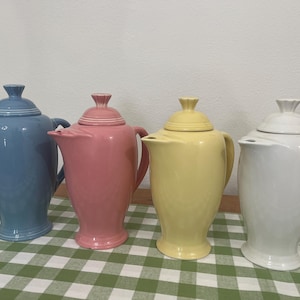 May include: Four ceramic teapots in pastel colors: light blue, pink, yellow, and white. Each teapot has a lid and a handle. The teapots are arranged on a green and white checkered tablecloth.