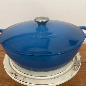 May include: A blue enameled cast iron Dutch oven with a lid and side handles. The lid has a silver knob. The pot is sitting on a white decorative plate. The background is a wooden surface.