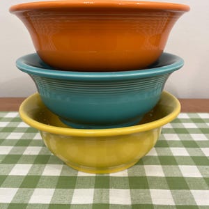 May include: Three colorful ceramic bowls stacked on top of each other. The top bowl is orange, the middle bowl is teal, and the bottom bowl is yellow. The bowls are sitting on a green and white checkered tablecloth.