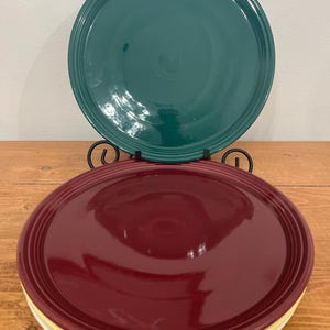 May include: A stack of three ceramic plates in teal, burgundy, and yellow. The teal plate is displayed on a black metal plate stand. The plates have a classic, simple design with a raised rim.