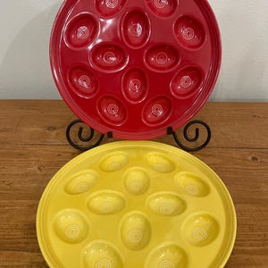 May include: Two ceramic serving platters, one red and one yellow, each with oval indentations. The red platter is on top, and the yellow platter is below. Both platters have a glossy finish and are designed for serving food.