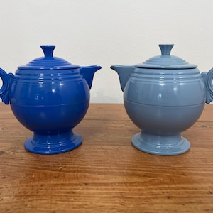 May include: Two ceramic teapots, one cobalt blue and the other a lighter blue, sit side-by-side. Each teapot has a round body, a lid with a small knob, a curved spout, and a circular handle. They are placed on a wooden surface.