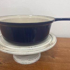 May include: A blue enameled cast iron pot with a white interior and a white handle. The pot is sitting on a white pedestal with decorative details.