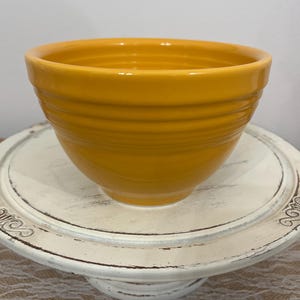 May include: A bright yellow ceramic mixing bowl with horizontal ridges. The bowl is sitting on a white, distressed, decorative cake stand. The stand has a round top and a pedestal base. The background is a neutral color.