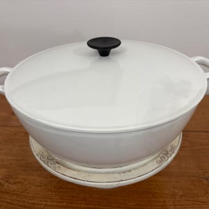 May include: A white enameled cast iron Dutch oven with a matching lid and black knob. The pot has two loop handles and sits on a decorative white and beige trivet. The cookware is on a wooden surface.