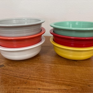 May include: Six ceramic bowls in various colors: gray, red, and white, and green, red, and yellow. The bowls are stacked in pairs, with a wide rim and a rounded base. The bowls are on a wooden surface.