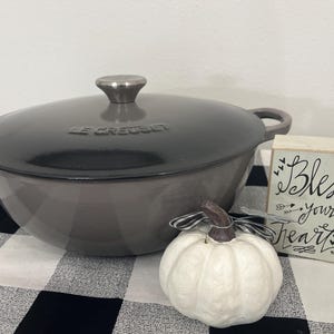 May include: A dark gray Le Creuset Dutch oven with a lid, a white pumpkin decoration, and a small wooden sign that reads "Bless your Heart" in black script. The items are on a black and white checkered tablecloth.
