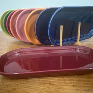 May include: A collection of seven oval ceramic trays in a spectrum of colors including lime green, red, pink, orange, dark blue, and a deep maroon tray positioned in the foreground. The trays feature a smooth, glossy finish and raised edges.