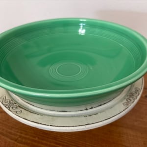 May include: A bright green ceramic bowl with a smooth, glossy finish and concentric ring detailing along the interior rim. The bowl is positioned on top of a white, circular, distressed-edge plate featuring decorative embossed scrollwork.