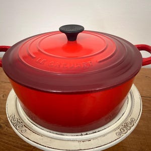 May include: A red Le Creuset Dutch oven with a black knob and side handles. The enameled cast iron pot sits on a white decorative stand. The lid has the brand name embossed on it.