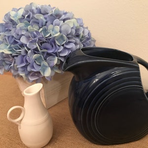 May include: A blue ceramic pitcher with a wide mouth and a curved handle. The pitcher has a textured, rippled design. The pitcher is sitting on a burlap surface next to a white ceramic pitcher and a bouquet of blue hydrangeas.