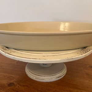 May include: A beige oval ceramic baking dish with handles sits on a white cake stand with a distressed finish.