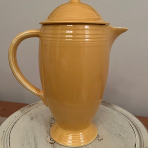 May include: A yellow ceramic coffee pot with a lid. The pot has a rounded body and a flared spout. The lid has a knob on top.