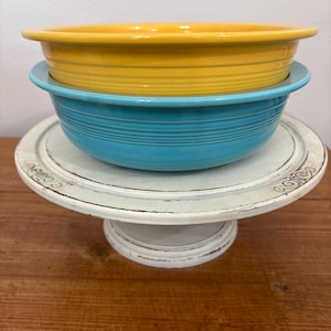 May include: A stack of two ceramic bowls, one yellow and one turquoise, resting on a white, distressed-look cake stand. The bowls have a ridged design. The cake stand has a decorative edge and a pedestal base.