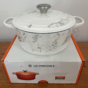 May include: White enameled cast iron Dutch oven with a marble pattern and a silver knob on the lid. The oven is sitting on a box with the Le Creuset logo and the words "Enameled Cast Iron" and "Lifetime" printed on it.