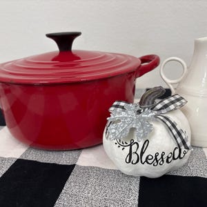 May include: A bright red enameled cast iron Dutch oven with a lid, a white ceramic pitcher with a handle, and a small white decorative pumpkin featuring a black and white gingham ribbon, a silver leaf accent, and the word Blessed in black script.