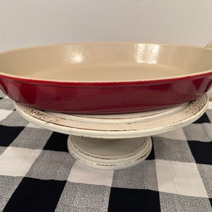 May include: A red oval baking dish with a cream-colored interior sits on a white cake stand. The stand has a distressed white finish and a decorative design around the top edge.