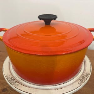 May include: An orange enameled cast iron Dutch oven with a matching lid and black handle. The pot has two side handles and sits on a white decorative stand. The cookware is a vibrant orange color.