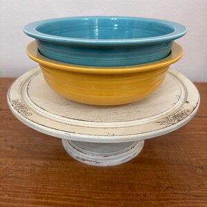 May include: Two ceramic bowls, one turquoise and one yellow, stacked on a white, distressed-look cake stand. The bowls have a simple, rounded design. The cake stand has a decorative edge and a pedestal base. The background is a plain white wall.
