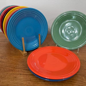 May include: A collection of colorful ceramic plates in various shades, including blue, red, yellow, and green. The plates are arranged on a wooden surface, with some stacked and others displayed individually. The plates have a classic, circular design.