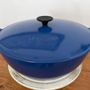 May include: A blue Le Creuset cast iron Dutch oven with a matching lid and black handle. The pot has two side handles and is sitting on a white plate. The Le Creuset logo is embossed on the lid.