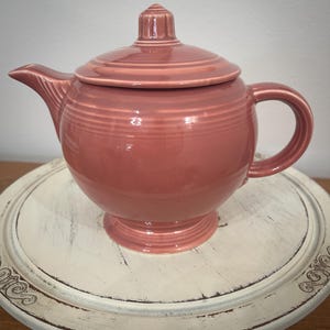 May include: A dusty rose-colored ceramic teapot with a lid, handle, and spout. The teapot has a round body and sits on a pedestal base. The lid has a small knob on top. The teapot is likely vintage or retro.