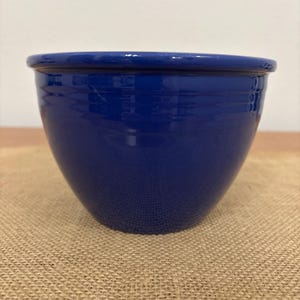 May include: A cobalt blue ceramic bowl with a rounded shape and a slightly flared rim. The bowl has a glossy finish and sits on a tan burlap surface. The bowl is empty and appears to be new.