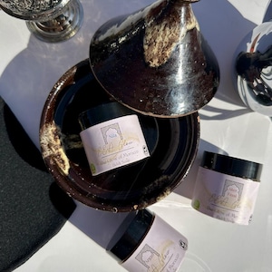 Puede incluir: Tres frascos de exfoliante corporal marroquí con diferentes etiquetas. Los frascos están sobre una superficie de cerámica marrón con fondo blanco. Las etiquetas están en inglés y árabe. Las etiquetas dicen "EEA Glow", "The Glow of Morocco", "Beldi Soap", "Nila", "Argan", "Aker Fassi".